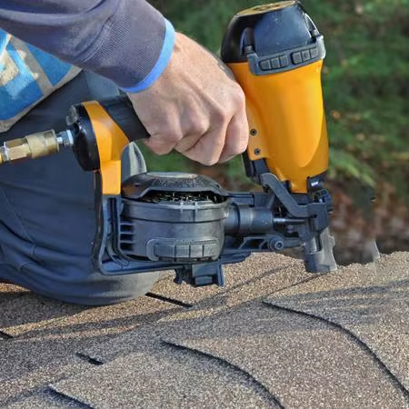 Worker using pneumatic coil nailer to drive big head coil nails into asphalt shingle roof during roofing installation